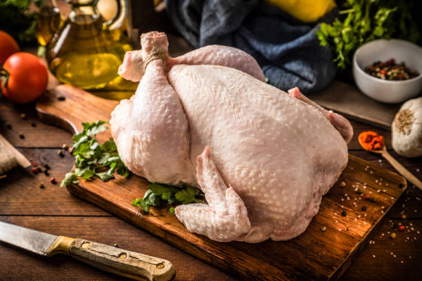 Fresh raw chicken on a rustic wooden table. Front view of a fresh raw chicken on a rustic wooden table. The chicken is on a cutting board and is surrounded by a kitchen knife and some ingredients such as a garlic clove, an olive oil bottle, some tomatoes, and parsley. raw food stock pictures, royalty-free photos & images