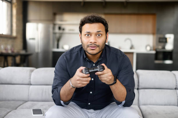 istock Involved in video game indian guy sits on the couch with a wireless joystick in hands 1323004680