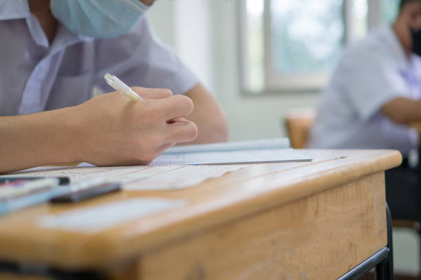 istock Hands of Asian students group learning and concentrate writing test in exam room wear facemask prevent COVID-19 coronavirus, Pupils Thai student uniform serious taking final examination on wood table 1323011685