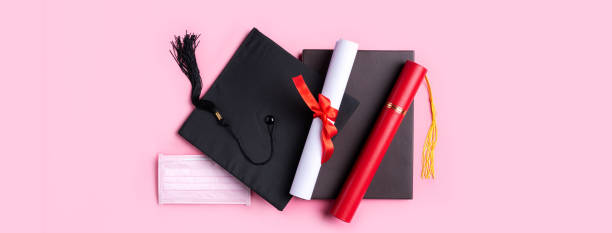 istock Graduation academic cap with diploma and mask isolated on pink table background. 1323018906