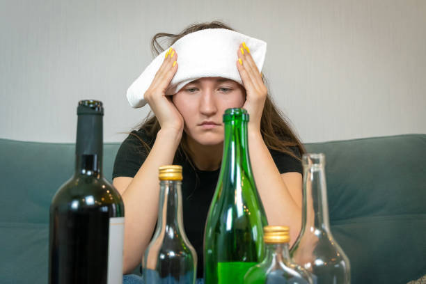 頭痛のした額に白いタオルを持つ若い女性は、アルコールの空のボトルの前に座っています。アルコールを飲んだ後の月曜日の朝、朝のコンセプト - feast day 写真 ストックフォトと画像