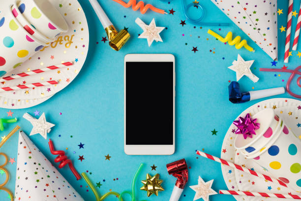 Top view photo of party table decoration smartphone in the middle paper plates cups hats pipes stars spiral tubes candles sequins and confetti on isolated blue wooden table background with copyspace vista superior foto de la decoración de la mesa de fiesta smartphone en las placas de papel del medio tazas sombreros tubos estrellas tubos espirales velas lentejuelas y confeti en el fondo de la mesa de madera azul aislado con copyspace