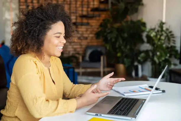 Side view at charming happy curly African-American young woman talking with online audience, using laptop, holding virtual meeting staying at home, dark skinned student girl involved seminar, webinar