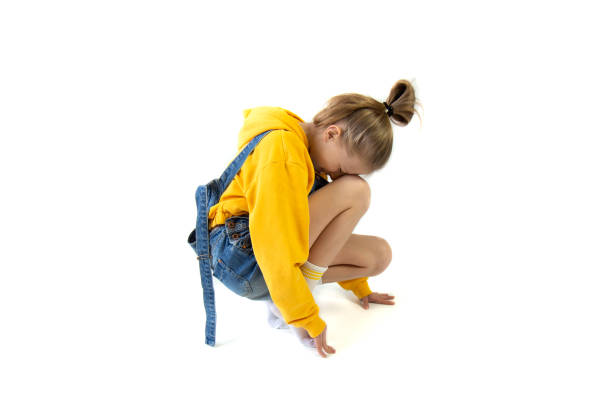 Teenage girl in denim overalls and in orange sweatshirt squatting on white background Beautiful teenage girl with a bun of hair in denim overalls and in orange sweatshirt squatting on white background. Girl is sad sad girl crouching stock pictures, royalty-free photos & images