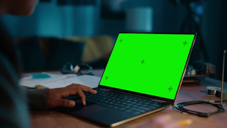 Young Teenage Multiethnic Girl Using Laptop Computer with Green Screen Chromakey Mock Up Display in a Dark Cozy Room at Home. She's Browsing Online. Doing School Homework Project.