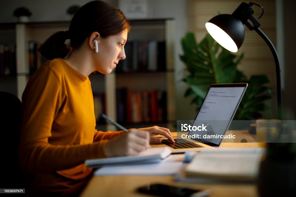 Teenage girl with bluetooth headphones studying late at home University Student Stock Photo