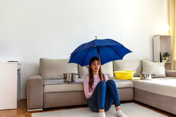 Photo of Worried woman sitting on the floor at home, holding umbrella under leaking ceiling.