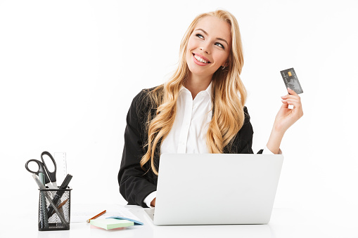 Photo of young businesswoman sitting at table and holding credit card while working on laptop isolated over white background in studio