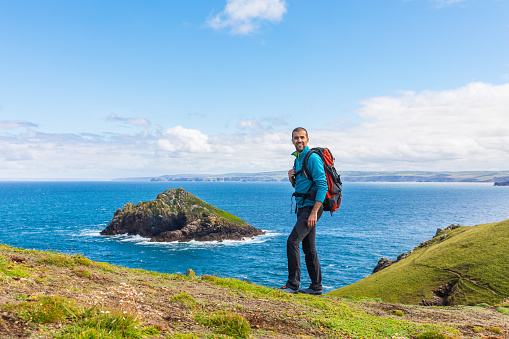 Man standing on a rock cliff enjoying the view - Young man with backpack and hiking clothes on the cliffs over seashore in Cornwall, UK - Travel, nature and wanderlust concepts.