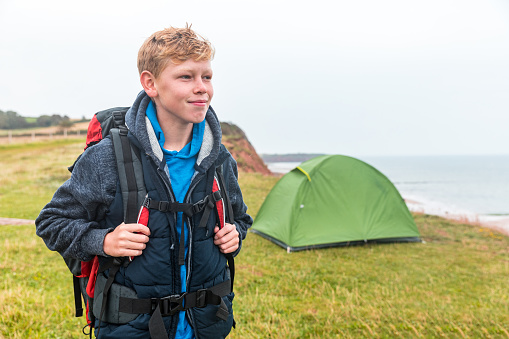 Happy boy portrait on hiking and camping trip - Young man wearing backpack and hiking gear posing in front of a tent on a seaside cliff - travel and adventure concepts
