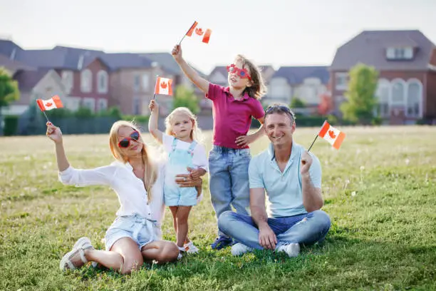 Photo of Happy Canada Day. Caucasian family with kids boy and girl sitting on ground grass in park and waving Canadian flags. Parents with kids children celebrating Canada Day on 1st of July outdoor