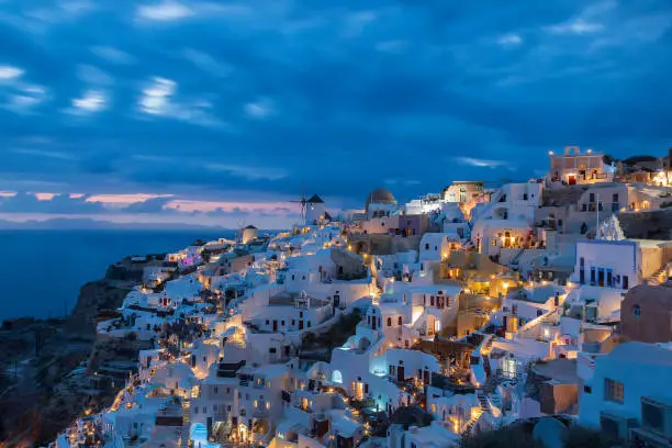 Evening view from the viewpoint of Oia town on Santorini in Greece after sunset. Beautiful clouds taken long time. The background is the sky at the blue hour. Lights are shining in white houses.