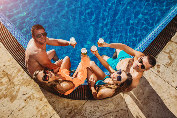 People drinking beer on the edge of a swimming pool stock photo. Youth and summer vacation concept. Group of happy friends cheering with beer on a sunny day by the pool. stock photo