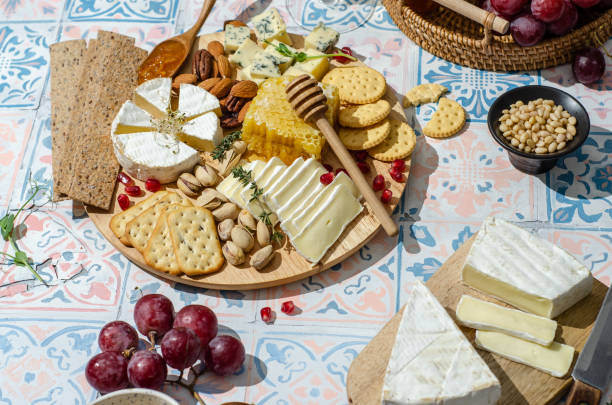 cheese plate with variety of appetizers on table. fresh brie cheese and camembert cheese whith crackers, pears, grapes, olives, rosemary, orange jam, orange jam, nuts, honeycomb and wine. - cheese emmental cheese swiss culture cutting board imagens e fotografias de stock