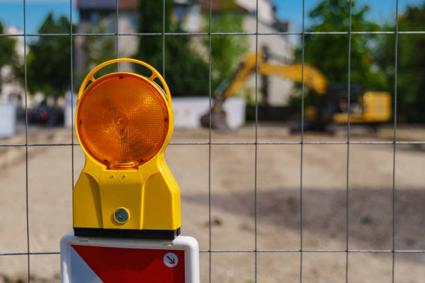 An orange warning light on the grating enclosing a construction site. Selective focus. Construction safety. Street barricade with warning signal lamp on a fence. Blur site background barricade stock pictures, royalty-free photos & images