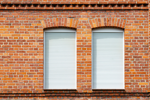Fragment of a brick wall of a building with two windows. The windows are covered with white blinds.