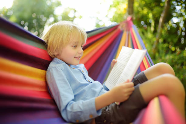 Cute little blond caucasian boy reading book and having fun with multicolored hammock in backyard or outdoor playground. Summer active leisure for kids. Child swinging and relaxing in hammock.