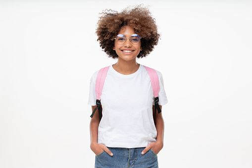 Retrato de una niña de escuela africana sonriente con camiseta blanca, gafas y mochila, aislada sobre fondo gris photo