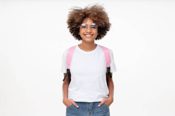 retrato de una niña de escuela africana sonriente con camiseta blanca, gafas y mochila, aislada sobre fondo gris - chica adolescente fotografías e imágenes de stock