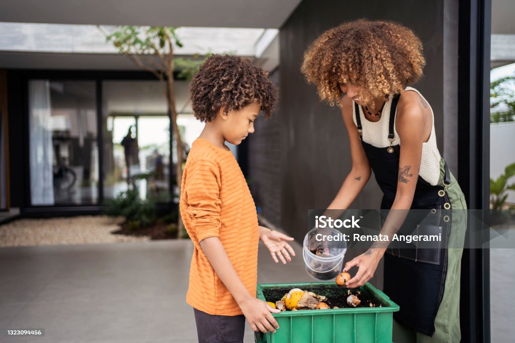 African american young woman and her younger brother making compost from leftovers. Sustainability concept. Young woman and her younger brother making compost from kitchen food leftovers into rich organic soil fertilizer. Compost Stock Photo
