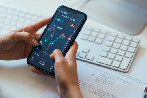 Hands of an unrecognisable woman with mobile phone on a desk, next to a keyboard, with a stock market chart on the screen