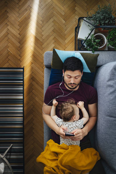 top vide of handsome dad using earphones and mobile phone while his little daughter is sleeping on him - babies and children audio imagens e fotografias de stock