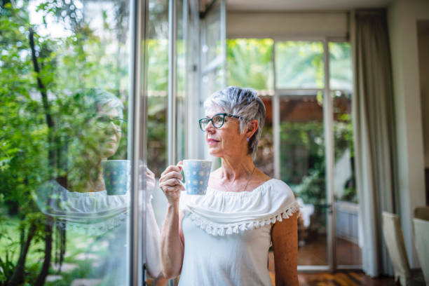 femme senior sereine avec du café regardant à travers la fenêtre - senior adult independence lifestyles home interior photos et images de collection