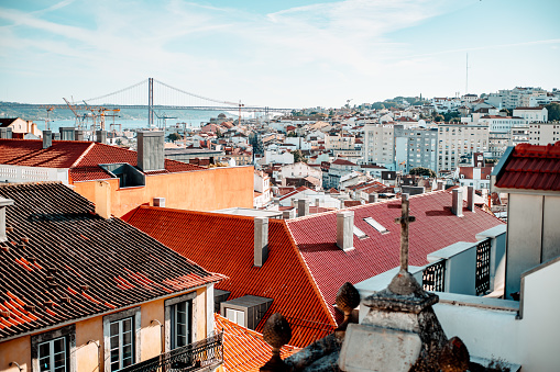 View of a sunny Lisbon cityscape from the roof; a Portuguese urban landscape with plenty of antique, old, and modern houses and a suspension bridge over Tejo river in the background