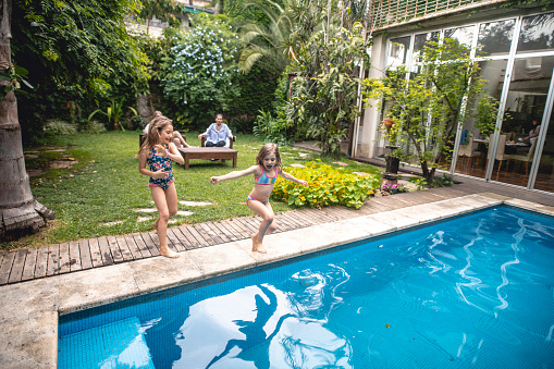 Young girl running past her sister to jump in family swimming pool while parents relax in background.