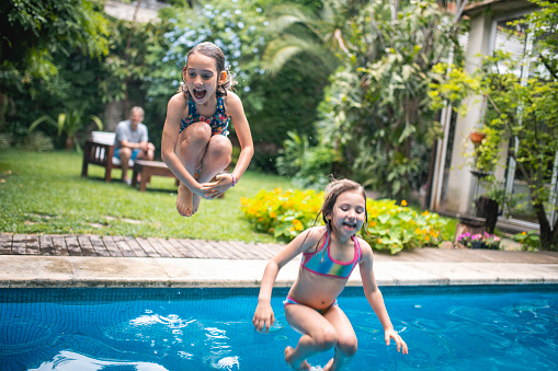 Laughing 6 and 8 year old sisters jumping together into swimming pool with family home exterior and backyard in background.