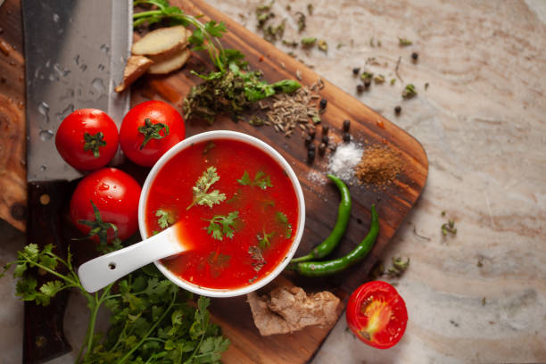 primo piano della zuppa di pomodoro indiana fatta in casa fresca e sana guarnita con foglie di coriandolo fresco e ingredienti ed erbe aromatiche, servita in una ciotola di ceramica bianca con un cucchiaio sopra la parte superiore in marmo. - zuppa di pomodoro foto e immagini stock