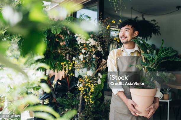 Confident Young Asian Male Florist Owner Of Small Business Flower Shop Holding Potted Plant Outside His Workplace He Is Looking Away With Smile Enjoying His Job To Be With The Flowers Small Business Concept Stock Photo - Download Image Now