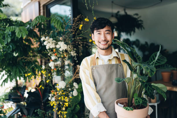 자신감 젊은 아시아 남성 꽃집, 중소 기업 꽃 가게의 소유자. 직장 밖에서 화분에 심은 식물을 들고 있습니다. 그는 미소를 지으며 카메라를 바라보고 있습니다. 꽃과 함께하는 그의 일을 즐기고 - i love my job 뉴스 사진 이미지