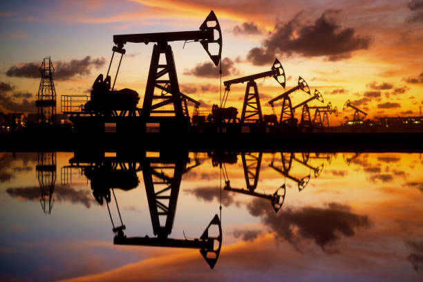 Oil Pumps And Rig At Sunset By The Sea Pump jack silhouette against a sunset sky with reflections in the water. oil field stock pictures, royalty-free photos & images