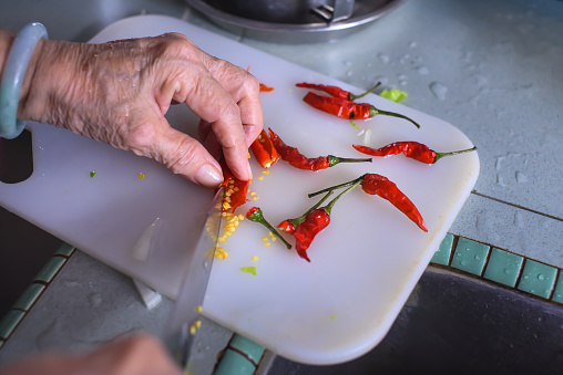 Close-up of senior woman cutting red chili peppers on a chopping board in home kitchen.