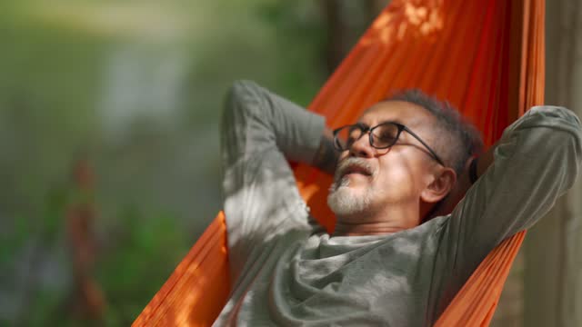 asian chinese senior man enjoying his afternoon lying down at hammock at riverside under bamboo trees