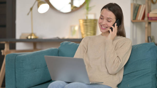 istock Young Woman with Laptop Talking on Smartphone on Sofa 1323162014