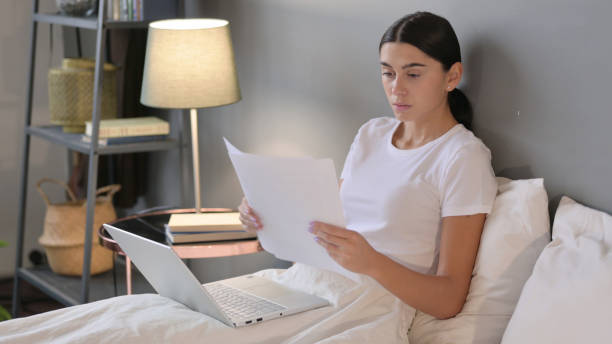 istock Latin Woman with Laptop Reading Documents in Bed 1323162121