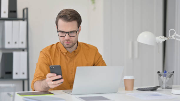 istock Young Man with Laptop using Smartphone in Office 1323163279