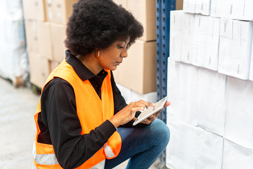 Woman worker using digital tablet while working in a warehouse. Female in reflective clothing sitting by a cargo rack and using tablet pc.