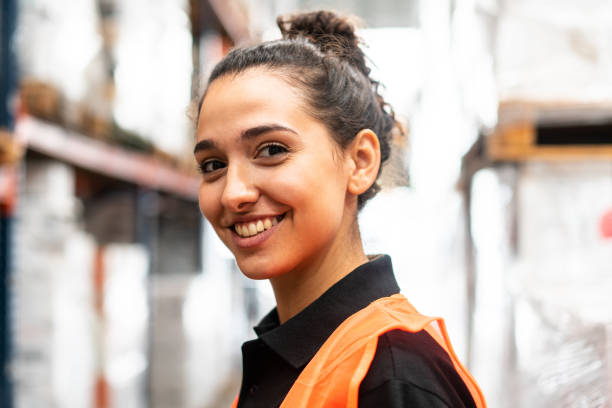 Close-up of a happy woman working in warehouse Close-up portrait of a happy young woman working in a distribution warehouse. Female worker looking at camera and smiling in the factory storage room. blue collar worker stock pictures, royalty-free photos & images