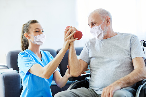 Doctor or nurse caregiver exercise with senior man, both wearing protective masks,  at home or nursing home
