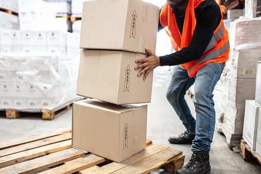 Cropped shot of a male worker in reflective clothing stacking boxes on a wooden pallet. Man working in distribution warehouse moving boxes.