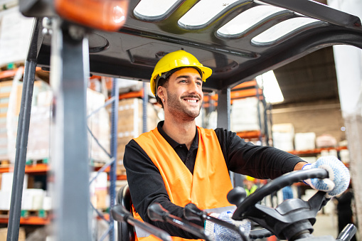 Young man operating a forklift loader in distribution warehouse. Male worker in hardhat and reflective vest driving a forklift.