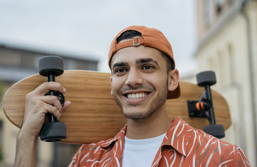 Close up portrait of young successful skater holding longboard, walking on the street, smiling. Happy Indian man wearing casual clothes posing for pictures outdoor