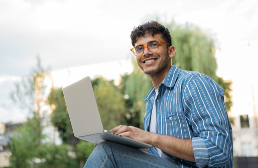 Portrait of handsome Indian copywriter using laptop computer, typing, working freelance project online, sitting outdoors. Smiling asian student studying, learning language, online education concept