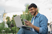 Indian man using laptop computer, mobile phone, working freelance project online, sitting outdoors. Successful business. Asian student studying, learning language, online education concept