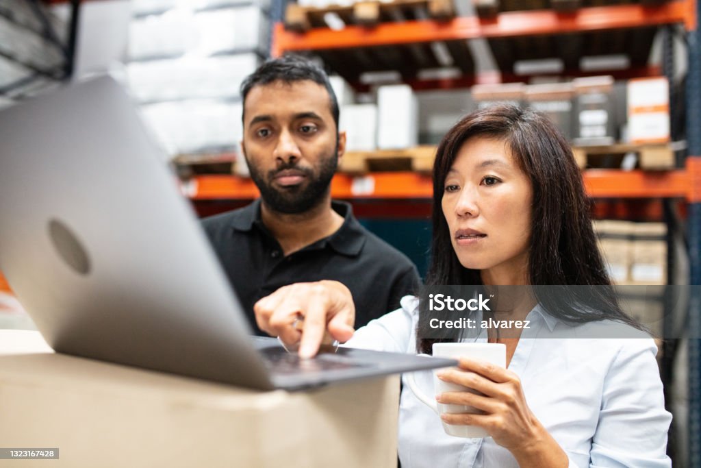 Warehouse staff working on a laptop Warehouse manager using laptop to share dispatch schedule with a male worker. Warehouse staff looking at laptop while working. Freight Transportation Stock Photo