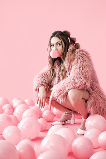 Funny hipster woman blowing bubble with chewing gum sitting in many pink balloons background