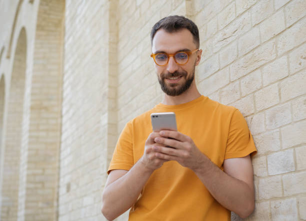 istock Handsome smiling man using mobile phone, communication online, chatting, watching video standing on the street. Young bearded hipster wearing stylish eyeglasses holding smartphone shopping online 1323169246